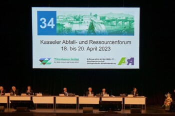 Klaus Wiemer (WI), Thomas Grundmann (ASA), Hendrik Engelkamp (EU), Peter Kurth (BDE), Patrick Hasenkamp (VKU), Henry Forster (bvse), Alexander Gosten (DGAW). Am Rednerpult: Susanne Lottermoser (BMUV) (von links) Blick auf das Podium mit sieben Experten und einer Expertin am Rednerpult, im Hintergrund eine Projektion: Kasseler Abfall- und Ressourcenforum 18. bis 20. April 2023